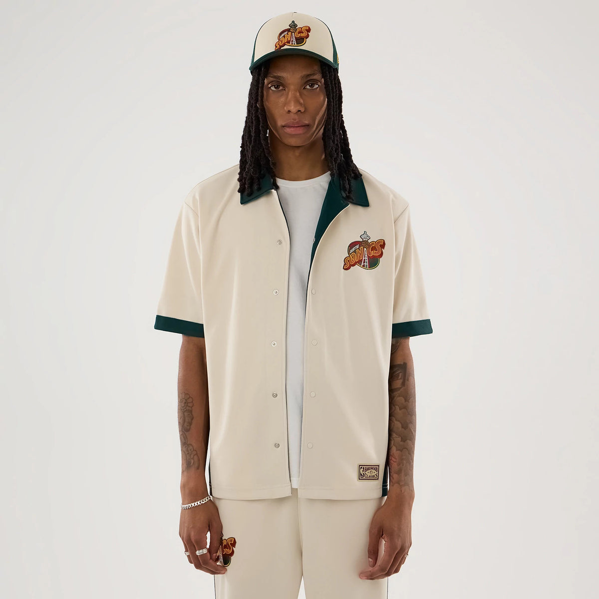 New Era Supersonics NBA Hardwood Classic Revere Beige Short Sleeve Shirt, front view on a model wearing a matching cap, featuring an embroidered Sonics logo on the left chest, green collar and sleeve trims, and buttoned front closure.
