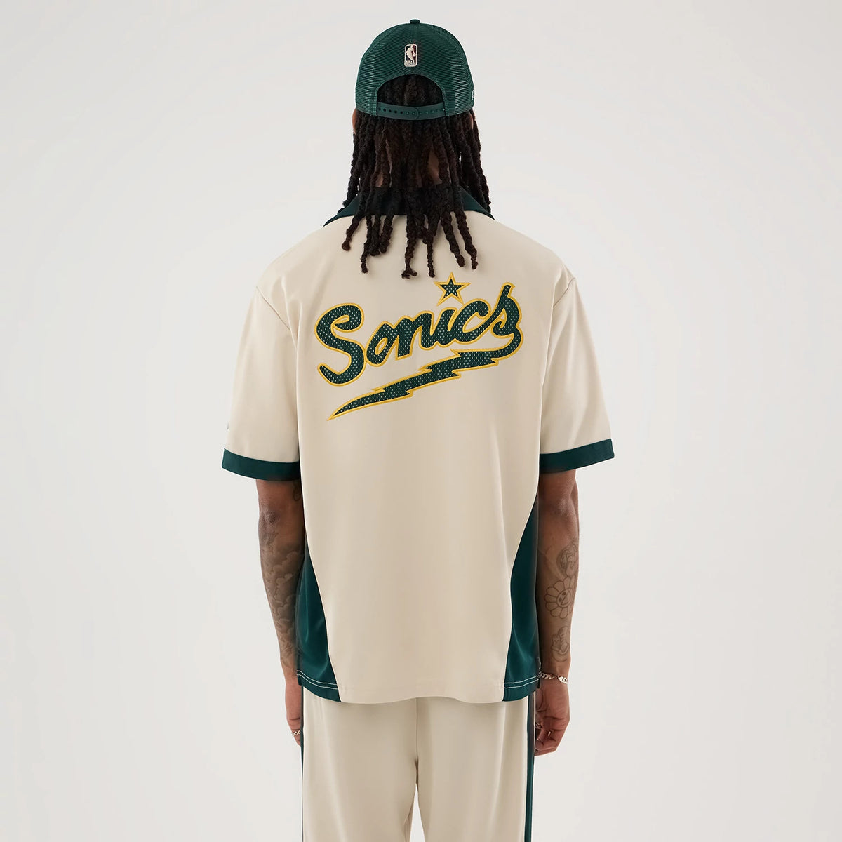 New Era Supersonics NBA Hardwood Classic Revere Beige Short Sleeve Shirt, back view on a model with a dark green cap, featuring large Sonics lettering with a star and lightning bolt on the back, and contrasting green trims on the sleeves.