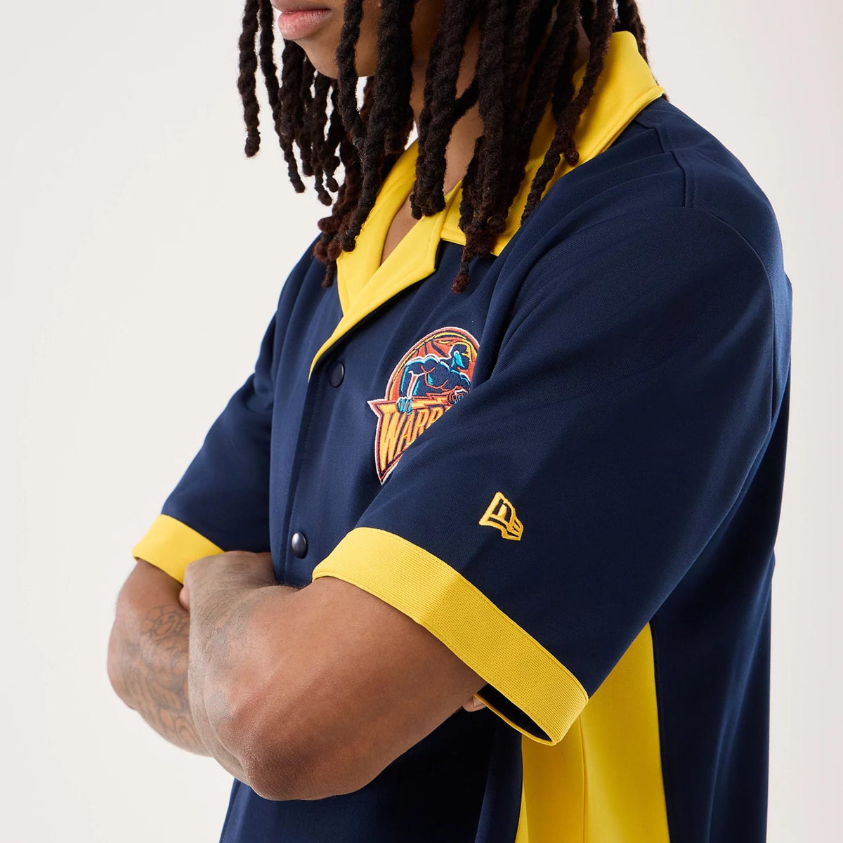 New Era Warriors NBA Hardwood Classic Revere Dark Blue Short Sleeve Shirt, side view on a model with the embroidered Warriors logo on the left chest, a yellow New Era flag logo on the sleeve, and contrasting yellow collar and sleeve trim.