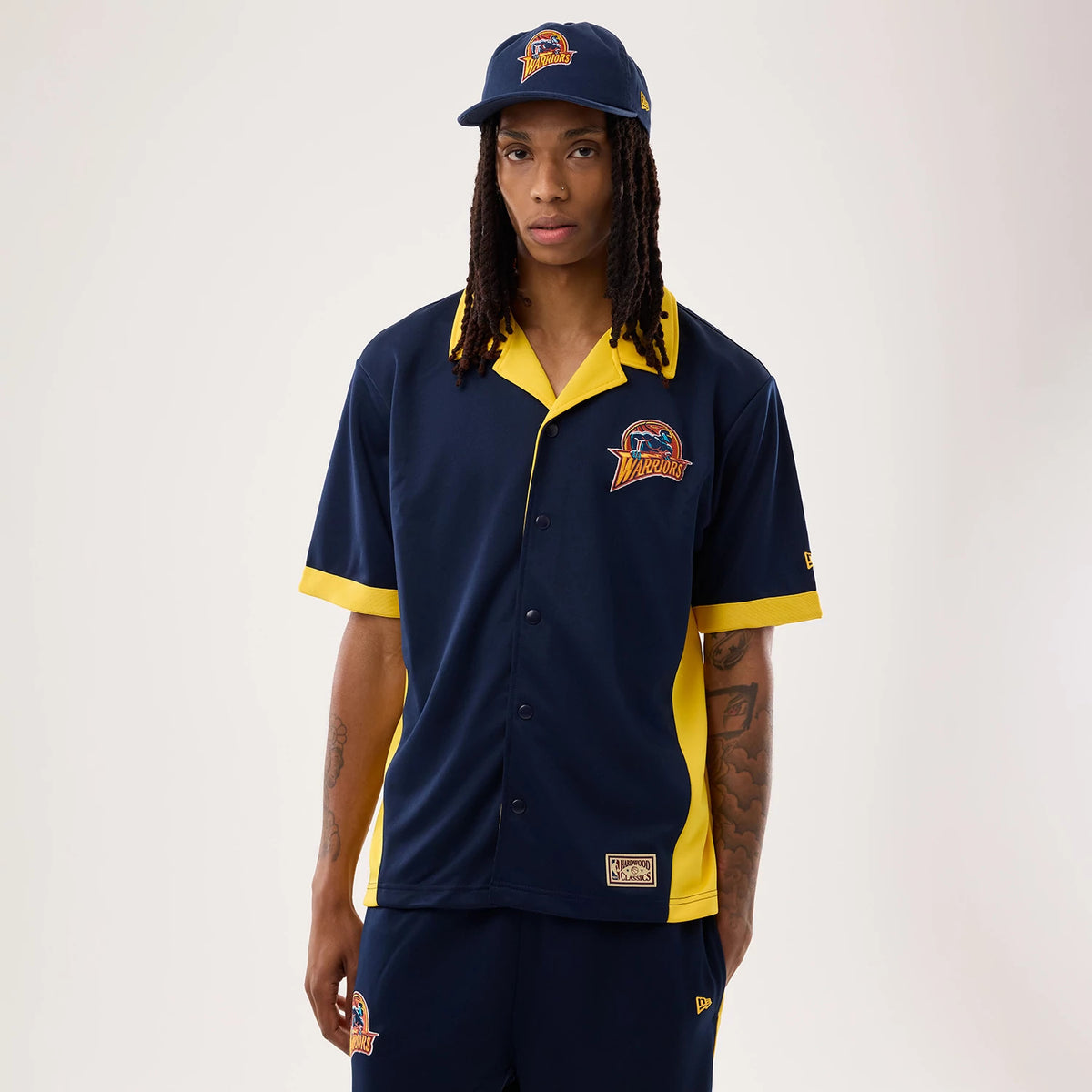 New Era Warriors NBA Hardwood Classic Revere Dark Blue Short Sleeve Shirt, front view on a model wearing a matching cap, featuring a Warriors logo embroidered on the left chest, yellow trim on the collar and sleeves, and a button-up design.