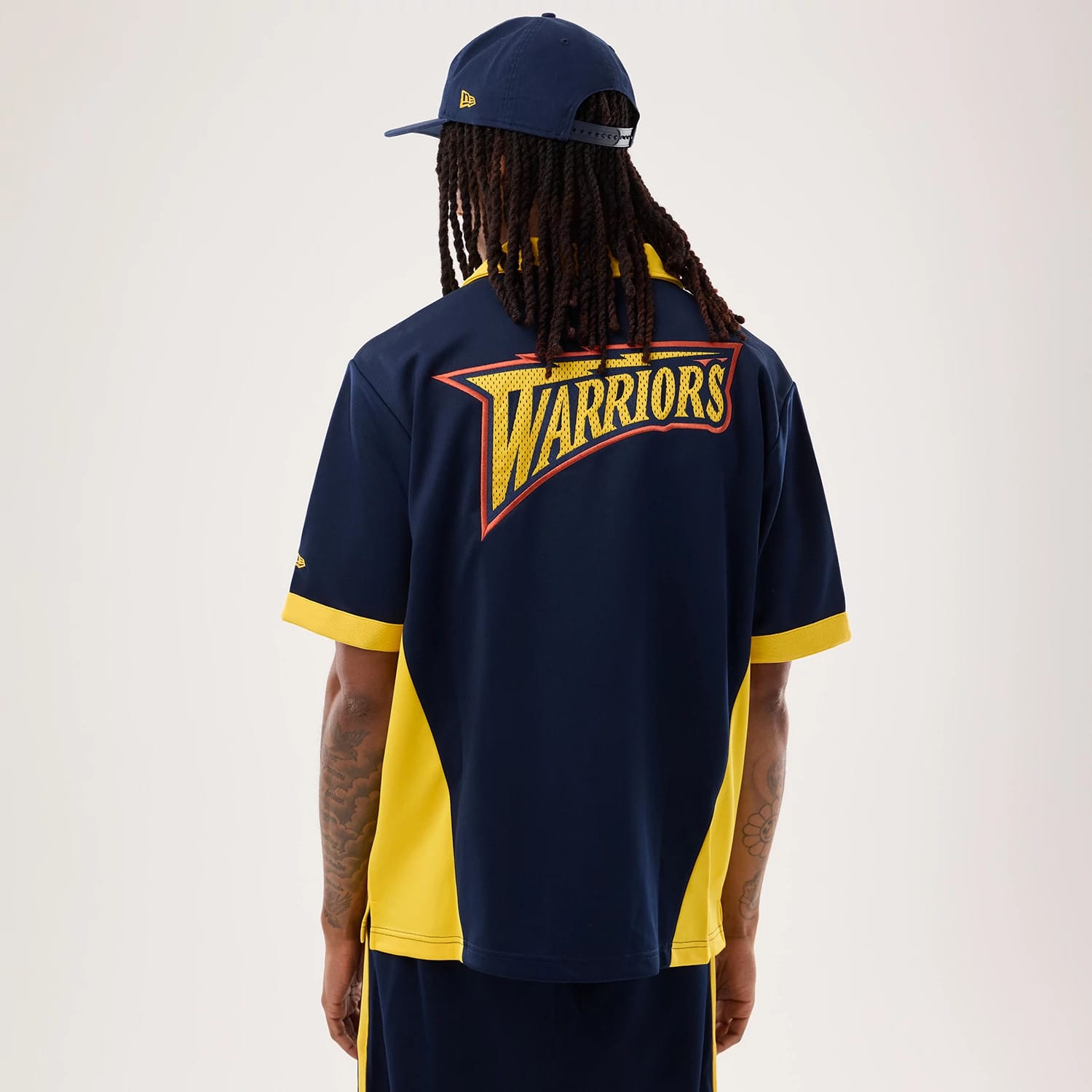 New Era Warriors NBA Hardwood Classic Revere Dark Blue Short Sleeve Shirt, back view on a model wearing a matching cap, featuring a large embroidered Warriors graphic across the upper back, yellow side panels, and contrasting yellow cuffs.