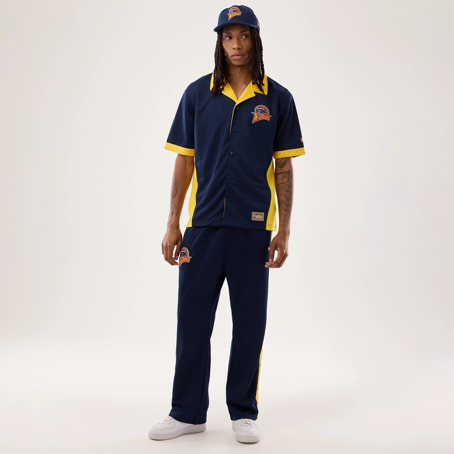 New Era Warriors NBA Hardwood Classic Revere Dark Blue Short Sleeve Shirt, front view on a model with a matching cap, showcasing a team logo on the left chest, contrasting yellow collar and trim, and button-front style.
