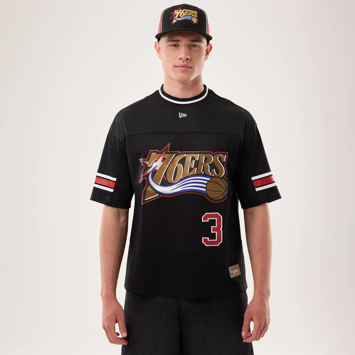 New Era Philadelphia 76ers NBA Hardwood Classic Mesh Oversized Black T-Shirt, front view on a model with a matching cap, featuring a large 76ers graphic across the chest, contrasting red and white striped sleeves.