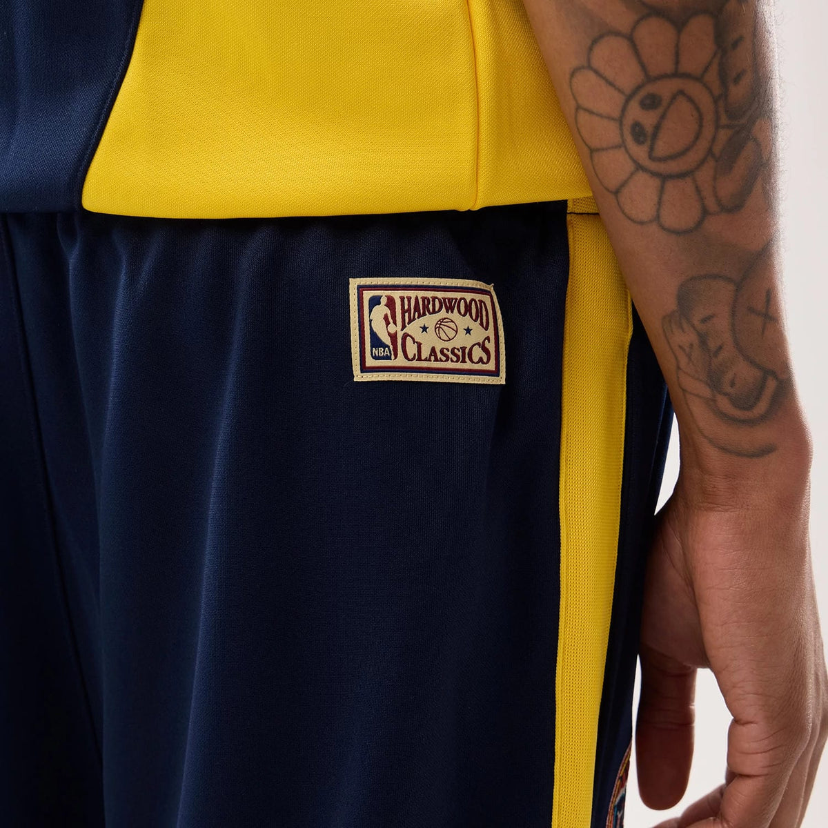 New Era Warriors NBA Hardwood Classic Straight Dark Blue Straight Leg Joggers, side view on a model with tattoos, showing the NBA Hardwood Classics patch on the upper left leg, dark blue fabric, and yellow side stripe detail.