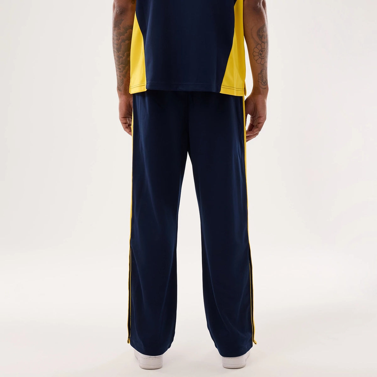 New Era Warriors NBA Hardwood Classic Straight Dark Blue Straight Leg Joggers, rear view on a model showing dark blue fabric with bright yellow side stripes along the legs, loose fit with elasticated waistband, paired with white trainers.