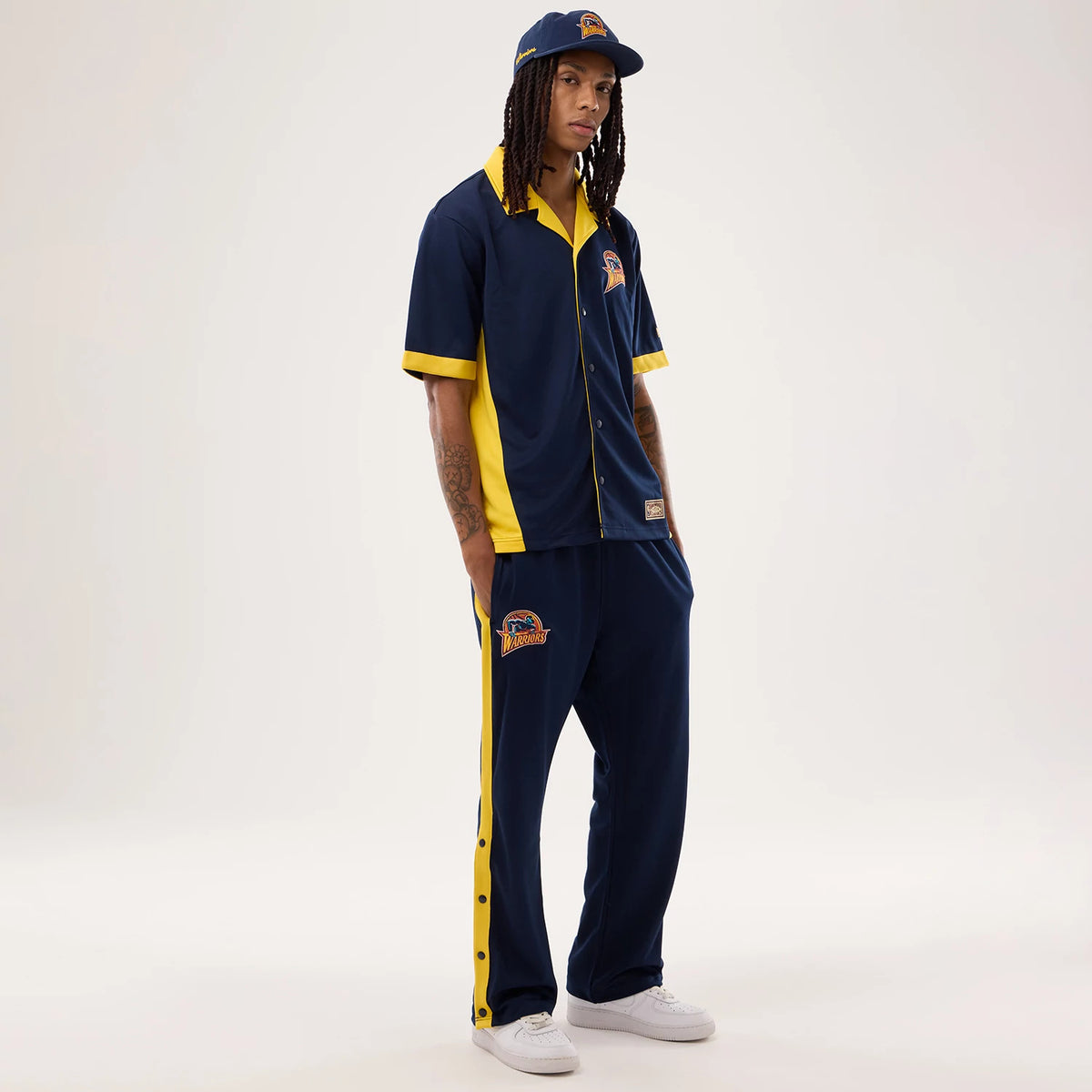 New Era Warriors NBA Hardwood Classic Straight Dark Blue Straight Leg Joggers, on-model view with yellow side stripes and a large Warriors embroidered patch on the left leg, paired with a matching shirt and white trainers.
