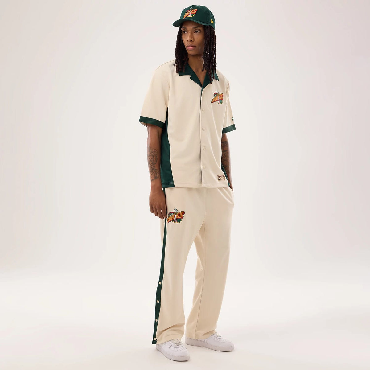 New Era Supersonics NBA Hardwood Classic Beige Straight Leg Joggers, on-model view featuring a green and orange retro Supersonics logo on the left thigh, dark green side stripes with white button accents, and elasticated waist with cream drawstrings.