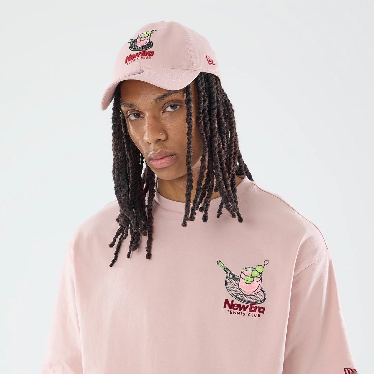 New Era Tennis Graphic Oversized Pastel Pink T-Shirt, front view on a model styled with matching cap, featuring a colourful embroidered graphic of a tennis racquet and cocktail on the left chest and a New Era logo on the sleeve.