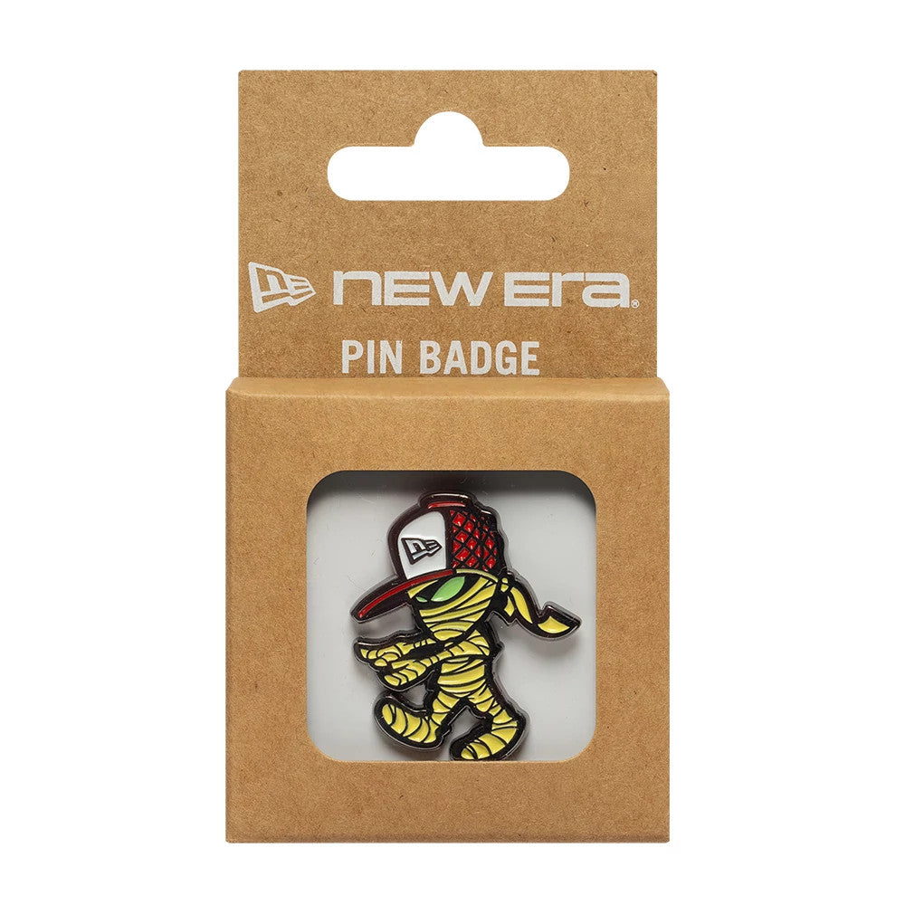 New Era Halloween Mummy Grey Pin Badge, front view showing a cartoon mummy character wrapped in yellow bandages, wearing a red baseball cap with a white New Era embroidered logo, displayed in a brown cardboard packaging with branding at the top.