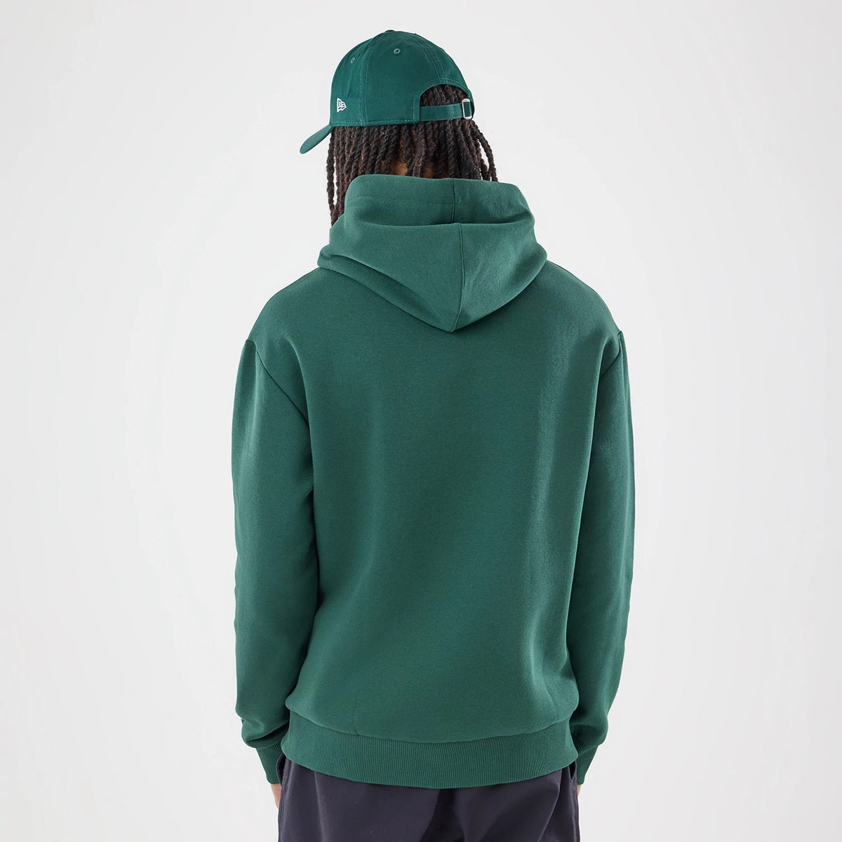 New Era Carlos Sainz Team Fifty5 Smooth Dark Green Oversized Pullover Hoodie, back view on a model featuring a generously sized hood and ribbed hem, styled with a matching dark green cap showcasing an embroidered New Era logo on the side.