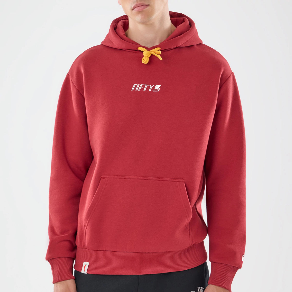 New Era Carlos Sainz Team Fifty5 Madrid Dark Red Oversized Pullover Hoodie, front view on a model showing the dark red hoodie with a yellow drawstring, embroidered Fifty5 logo on the chest and a ribbed hem, paired with black trousers.