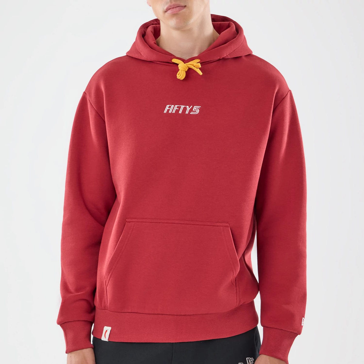 New Era Carlos Sainz Team Fifty5 Madrid Dark Red Oversized Pullover Hoodie, front view on a model showing the dark red hoodie with a yellow drawstring, embroidered Fifty5 logo on the chest and a ribbed hem, paired with black trousers.
