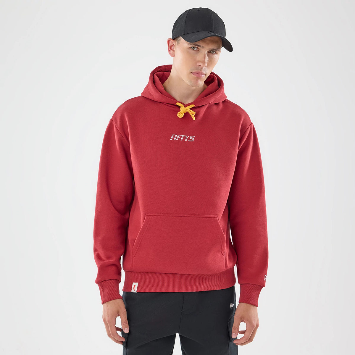 New Era Carlos Sainz Team Fifty5 Madrid Dark Red Oversized Pullover Hoodie, front view on-model featuring a yellow drawstring, embroidered Fifty5 logo across the chest, and a kangaroo pocket, styled with black trousers and a cap for a casual look.