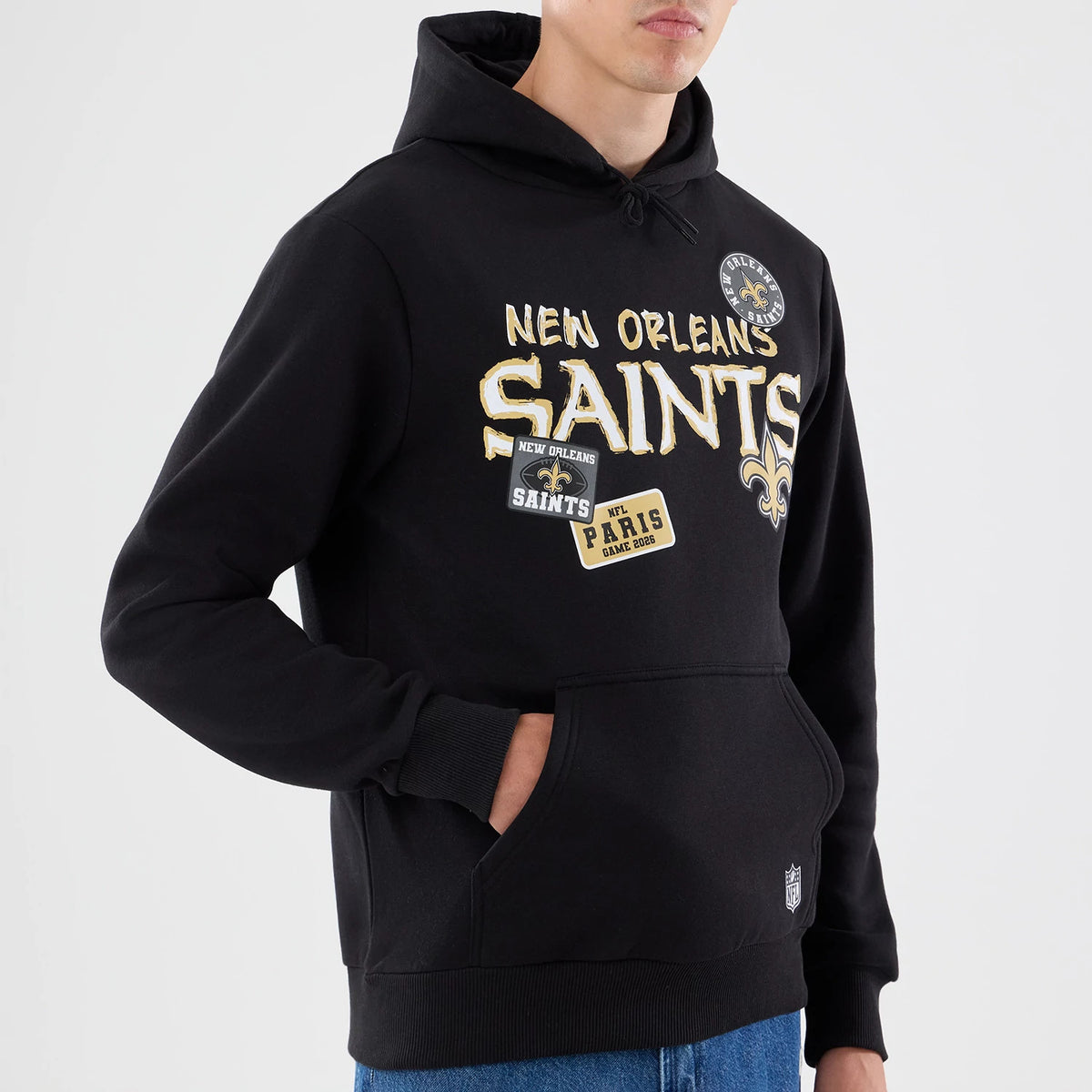 New Era New Orleans Saints NFL Patch Black Pullover Hoodie, front view featuring embroidered New Orleans Saints lettering across the chest, multiple NFL-themed patches, a large front pocket, and an embroidered NFL logo near the waistband on a model.