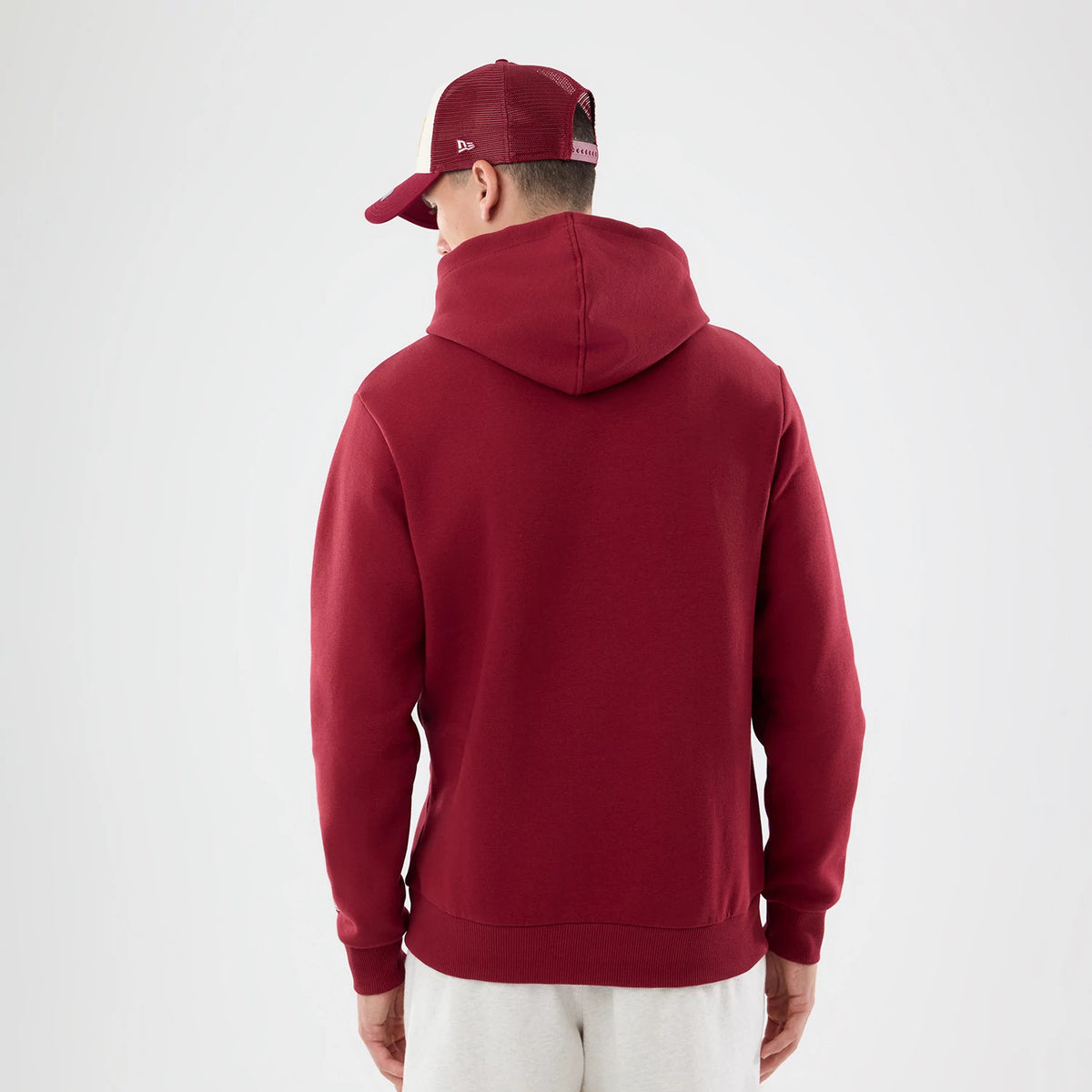 New Era Washington Commanders NFL Patch Dark Red Pullover Hoodie, back view on a model styled with a matching cap, showcasing the solid dark red fabric with no logos, a large hood draped over the shoulders and ribbed cuffs at the wrists.