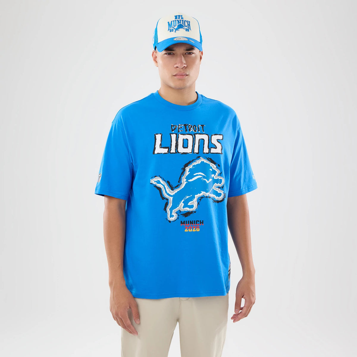 New Era Detroit Lions NFL Graphic Slogan Blue Oversized T-Shirt, front view on-model featuring a printed Detroit Lions graphic across the chest, an oversized lion logo below, with additional Munich 2023 lettering, styled with a coordinating cap.
