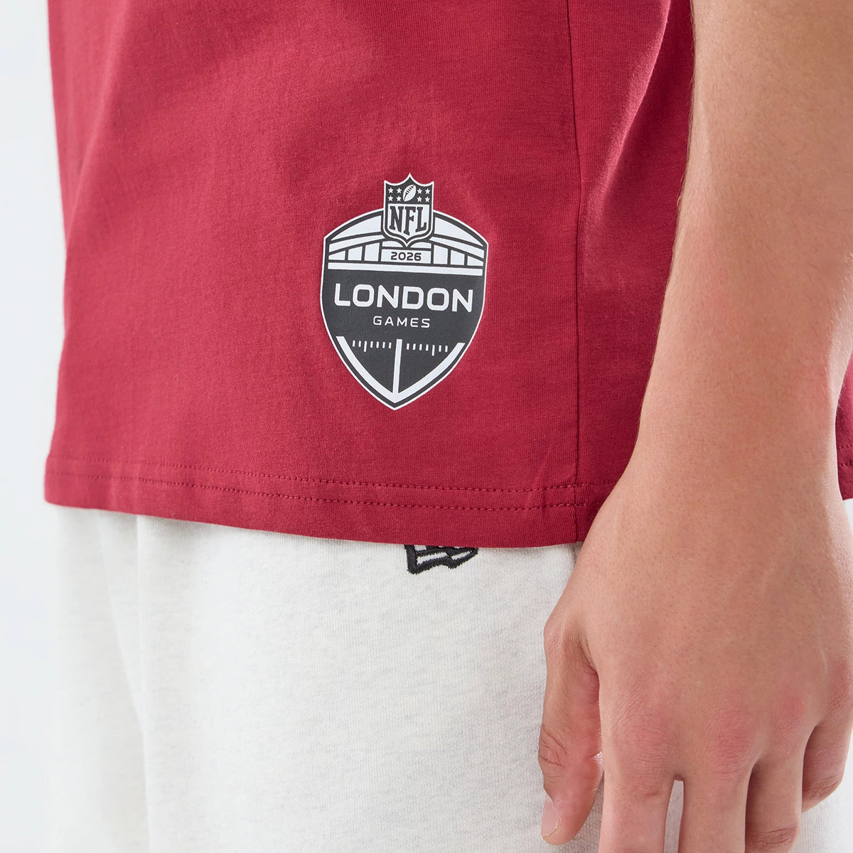 New Era Washington Commanders NFL Graphic Slogan Dark Red Oversized T-Shirt, side view highlighting a printed NFL 2026 London Games badge near the hem, paired with light shorts showing an embroidered New Era logo just below the waistband.