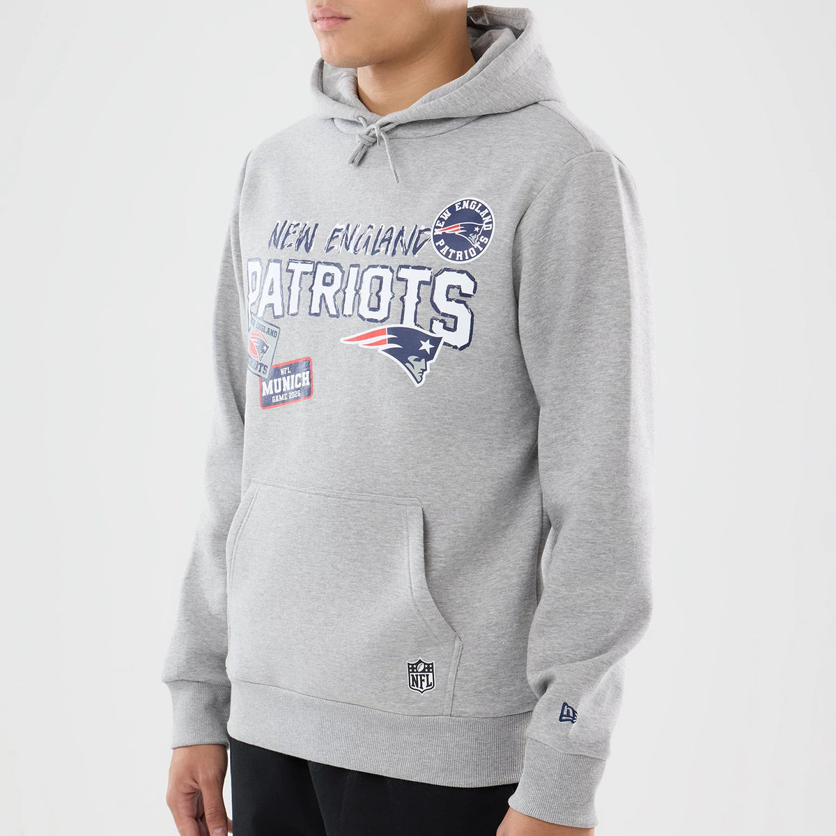 New Era New England Patriots NFL International Games 2026 Grey Pullover Hoodie, front view on a model showing the Patriots lettering across the chest, embroidered team logo beneath, round team patch on the shoulder.