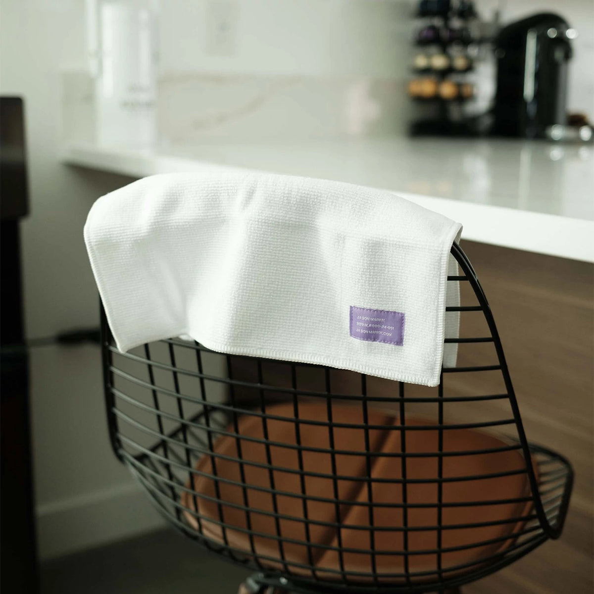 New Era Jason Markk® Absorb Premium Microfiber Towel, draped over a wire chair with a visible purple label on a white textured fabric, against a kitchen background featuring a countertop and coffee machine.