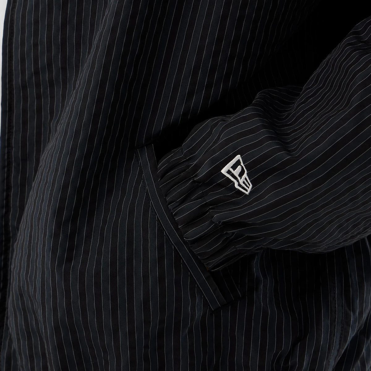 New Era Street Class Navy Jacket, close-up view showing the black and white pinstripe pattern on the sleeve, with a raised New Era logo embroidered near the cuff and a side pocket detail beneath.