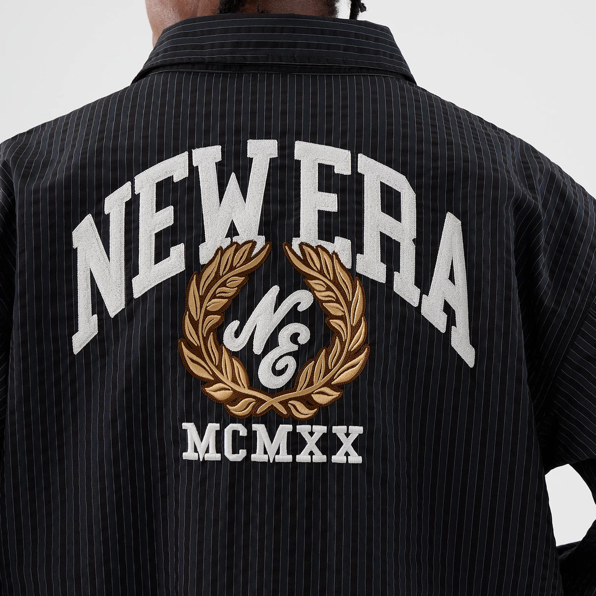 New Era Street Class Navy Jacket, back view showing large white embroidered New Era lettering across the upper back, a brown and gold embroidered wreath with "NE" inside, and "MCMXX" below on the dark navy pinstriped fabric.