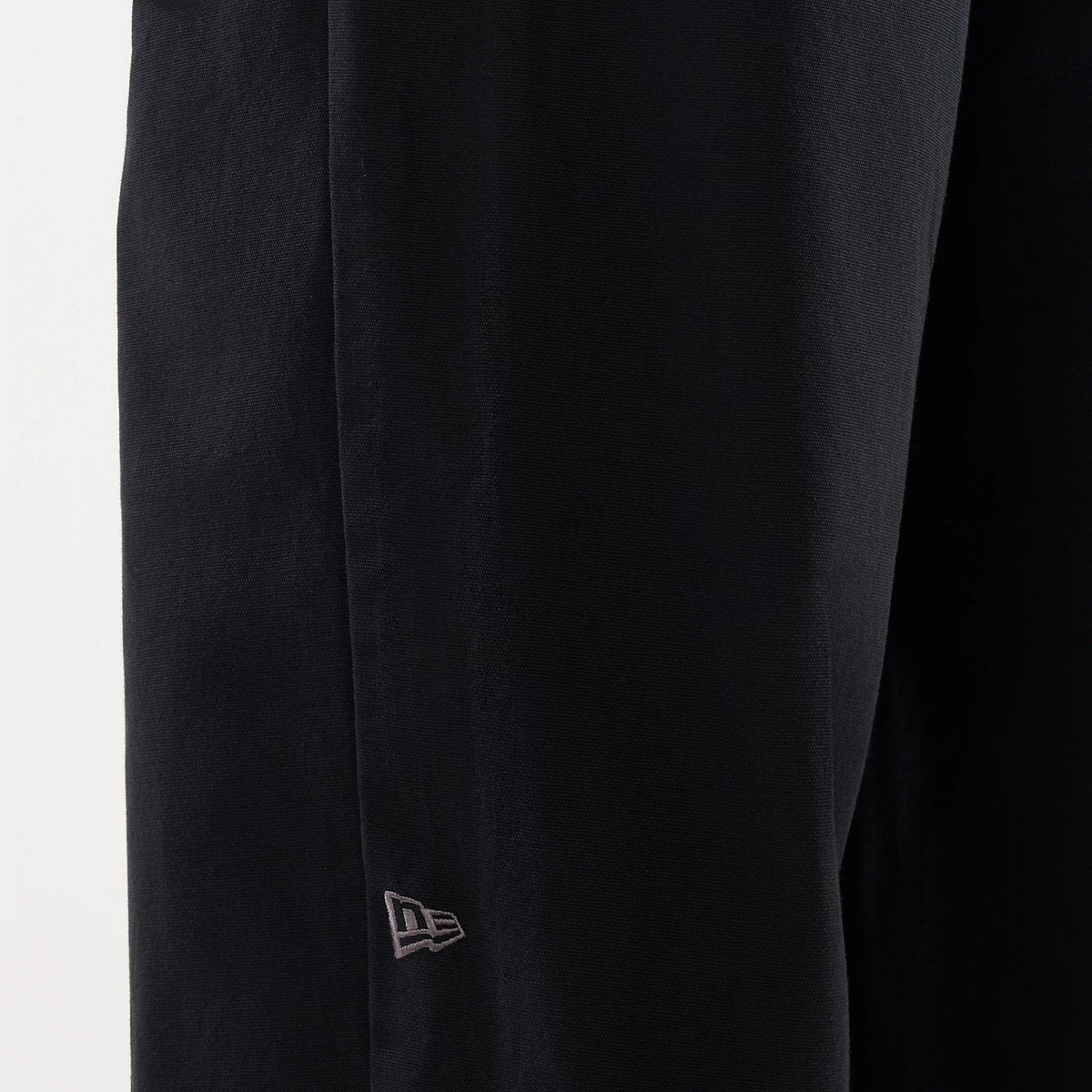 New Era Black Balloon Leg Trousers, close-up showing a subtle New Era flag logo embroidered near the seam on the smooth black fabric, emphasizing the clean silhouette and relaxed fit style.