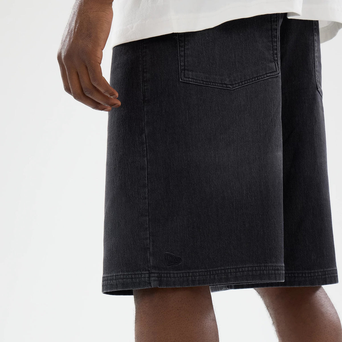 New Era Washed Black Bermuda Shorts, side view showing the faded black fabric with a debossed New Era logo above the hem, a patch pocket on the back, and straight leg openings below the knee.
