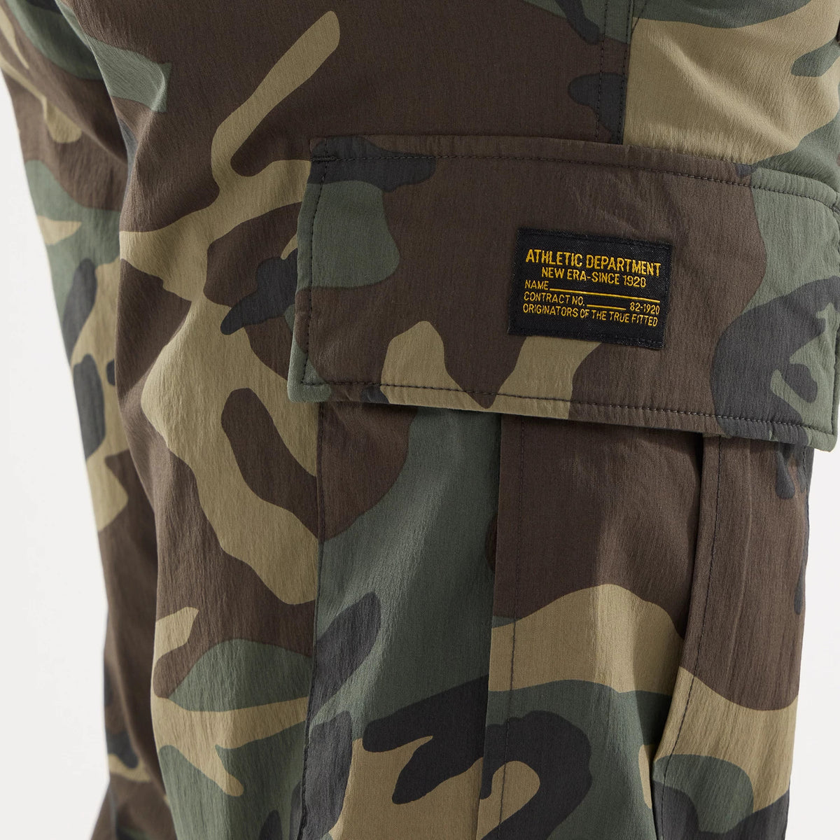 New Era Originators Woodland Camo Cargo Trousers, close-up showing the camo pattern on the cargo pocket with a black and yellow woven label reading “Athletic Department New Era Since 1920 Originators of the True Fitted”.