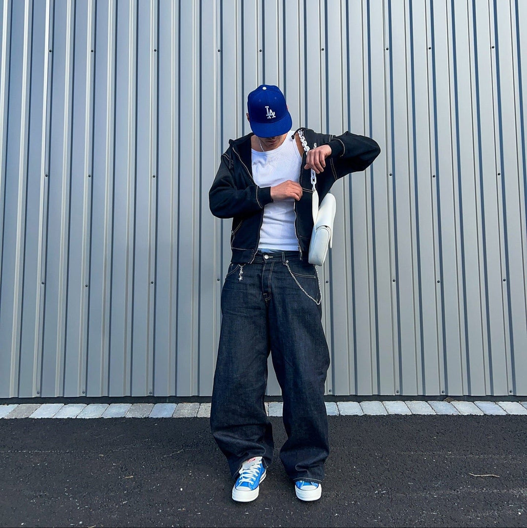 Person wearing a blue cap, black jacket, white shirt, dark jeans, and blue sneakers standing against a gray wall.