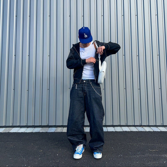 Person wearing a blue cap, black jacket, white shirt, dark jeans, and blue sneakers standing against a gray wall.
