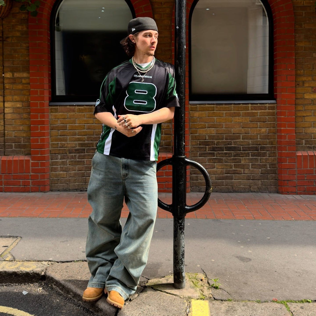 Person wearing a green and black sports jersey with the number 8, and a New Era Cap, standing on a street corner.