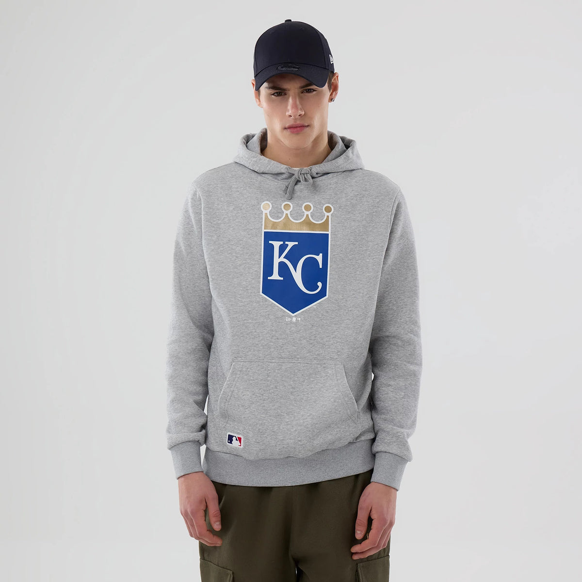 New Era Kansas City Royals MLB Grey Pullover Hoodie, front view showing a large centrally placed blue and gold team logo on the chest, with a drawstring hood and ribbed cuffs, on-model with khaki trousers and a navy cap.