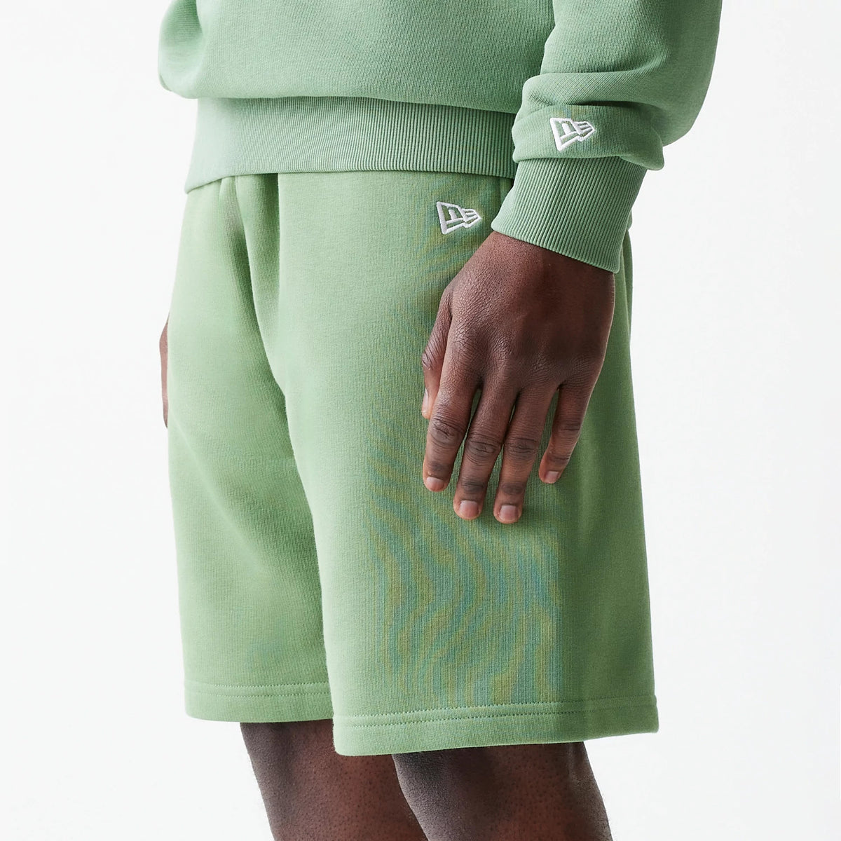 New Era Essential Green Oversized Shorts, side view showing the relaxed fit with a raised New Era logo on the left thigh, complemented by a matching green sweatshirt with a raised logo on the cuff.