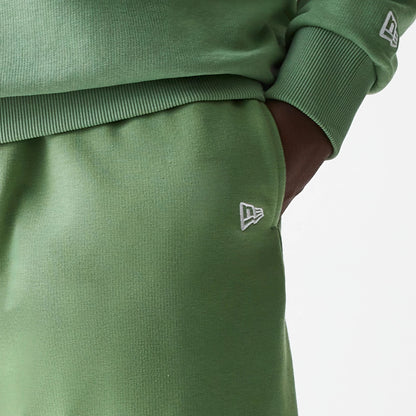 New Era Essential Green Oversized Shorts, on-model view showing the green fabric with a raised white New Era logo on the left thigh and ribbed waistband, paired with a matching green top featuring a similar logo on the cuff.