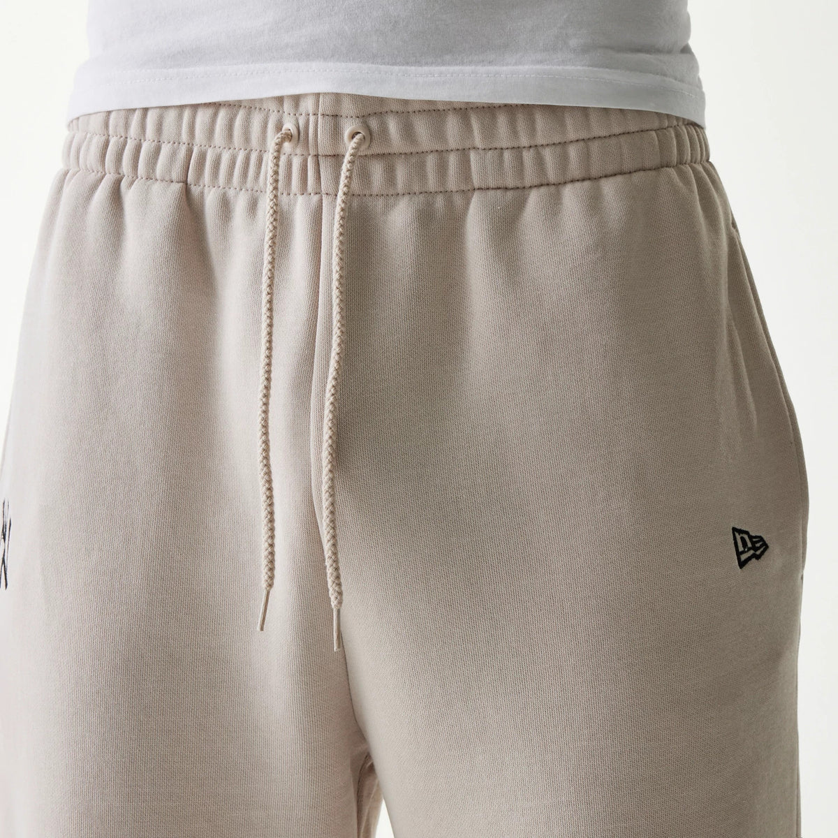 New Era New York Yankees MLB League Essential Cream Joggers, front view showing cream drawstrings, elastic waistband, and an embroidered New Era flag logo on the right leg, worn with a white T-shirt.