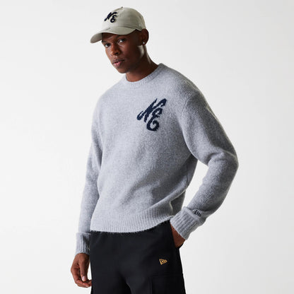 New Era Knit Grey Oversized Knit Sweater, on a model paired with black trousers and a cream cap, showcasing the navy NE logo embroidered on the chest and the New Era flag logo on the left cuff.