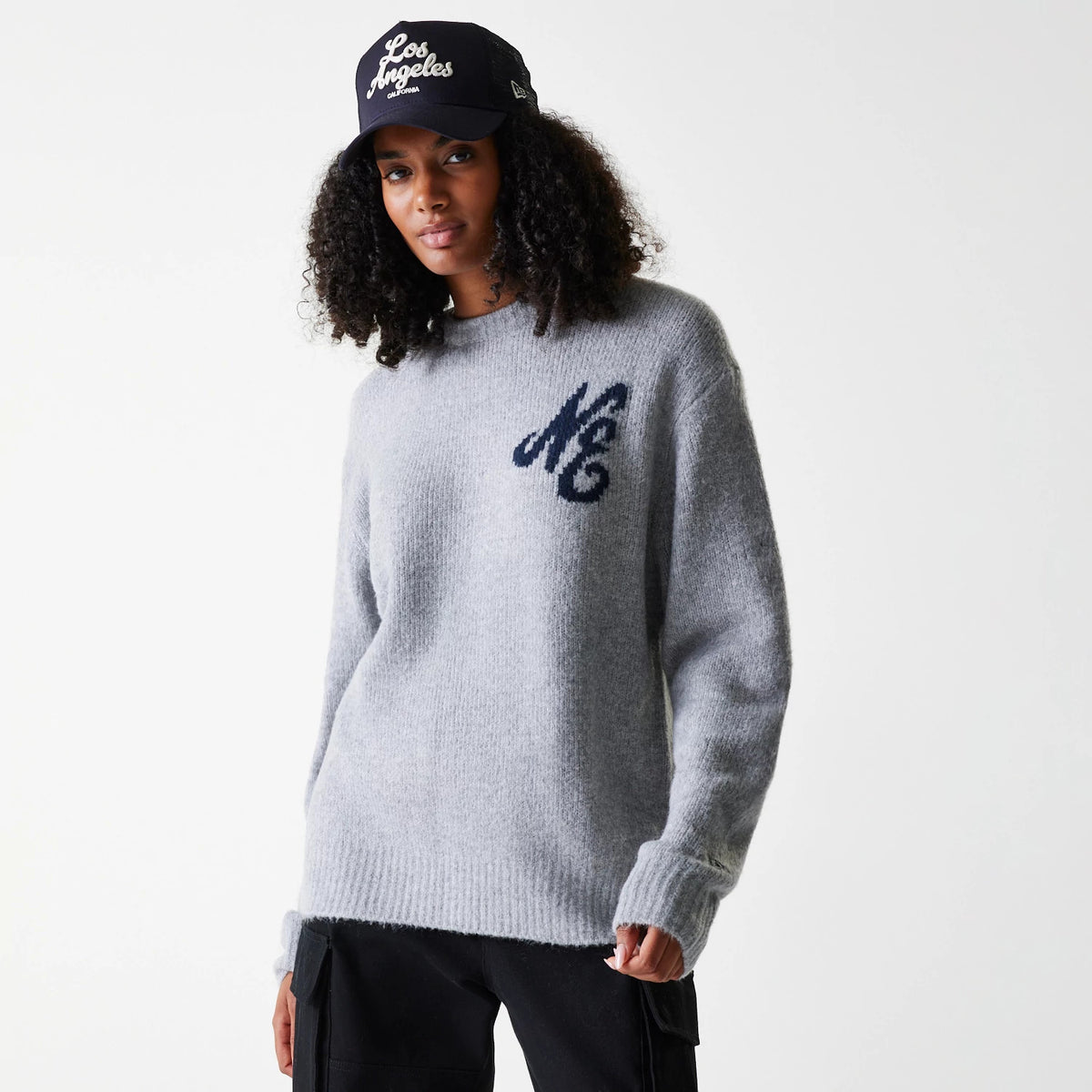 New Era Knit Grey Oversized Knit Sweater, on a model styled with a black cap, featuring an embroidered MC logo on the left chest and a ribbed hem and cuffs, highlighting a casual and relaxed fit.