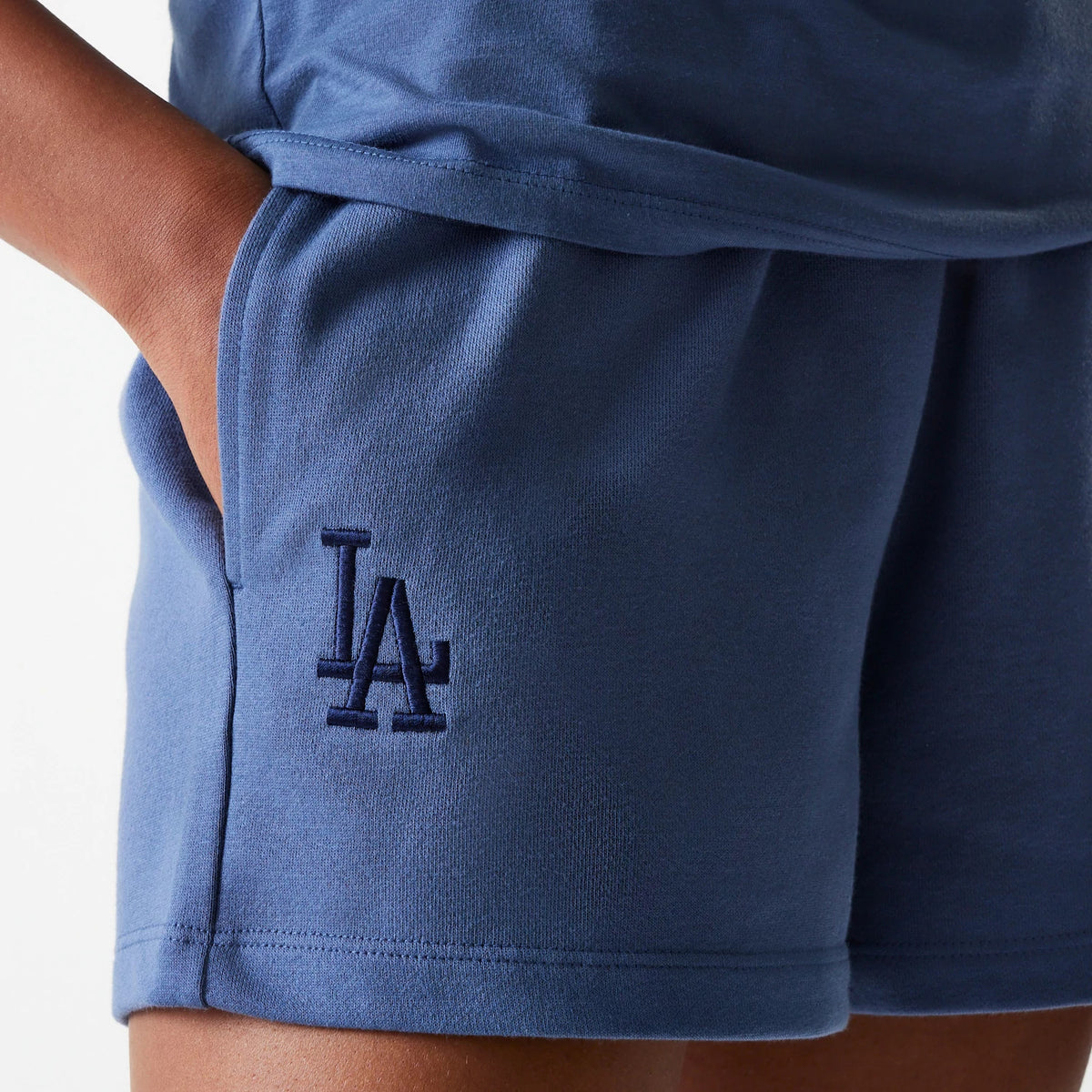 New Era LA Dodgers Womens MLB Midi League Essential Dark Blue Shorts, side view showing the dark blue fabric with an embroidered LA logo on the left lower leg, a hand placed in the side pocket of the mid-rise waistband.