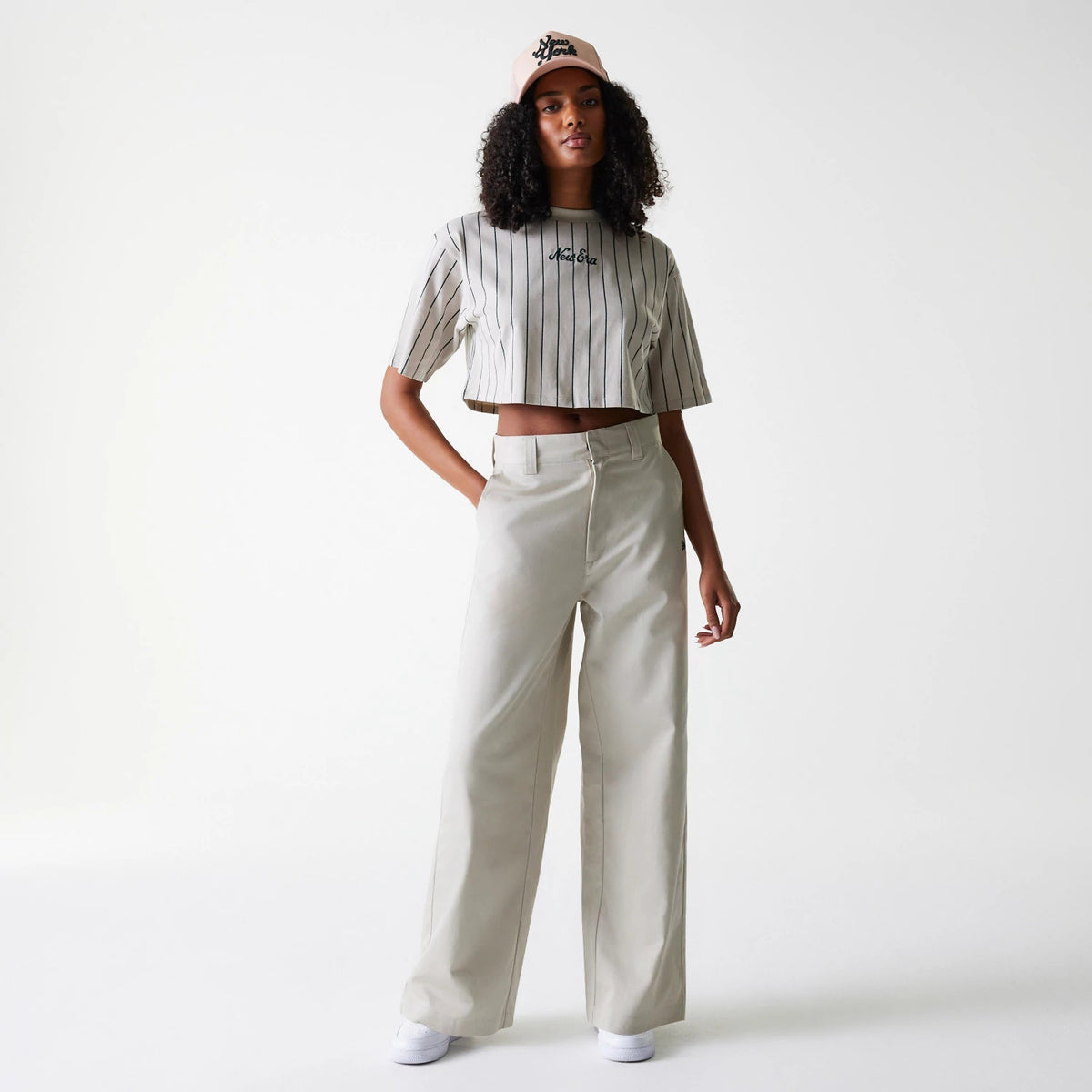 Womens New Era Light Beige Wide Leg Chino Trousers, on-model showing full-length trousers with loose fit and clean lines, paired with a striped crop top and light pink New Era cap with script logo.