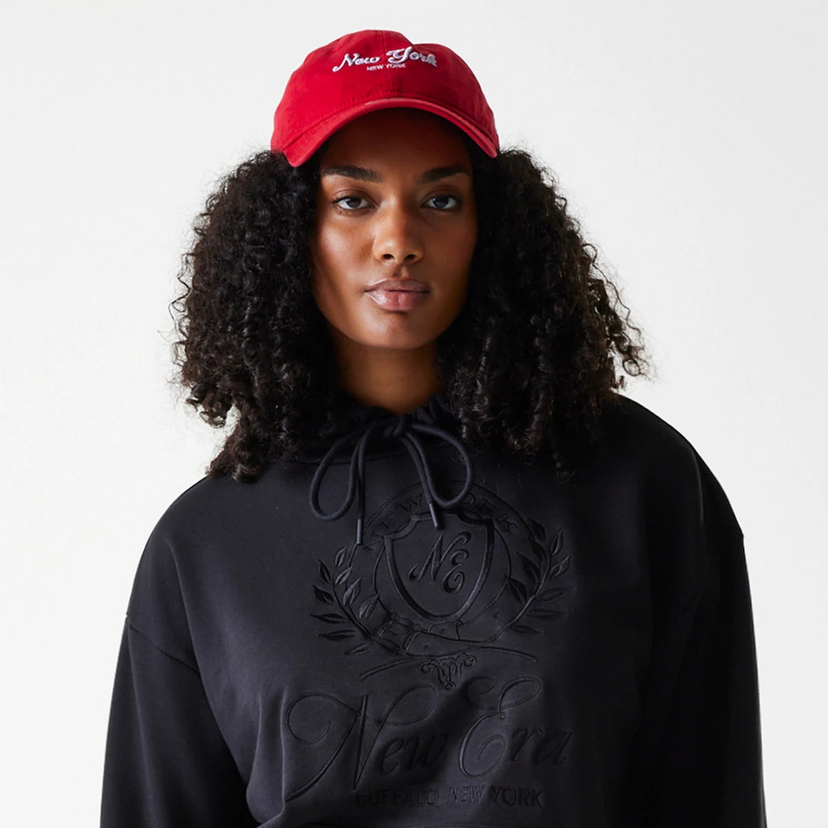 Womens New Era Crest Black Pullover Hoodie, on-model view showing the black hoodie with a large embossed New Era logo on the chest, drawstrings at the neckline, and worn under a red cap featuring a white embroidered logo.