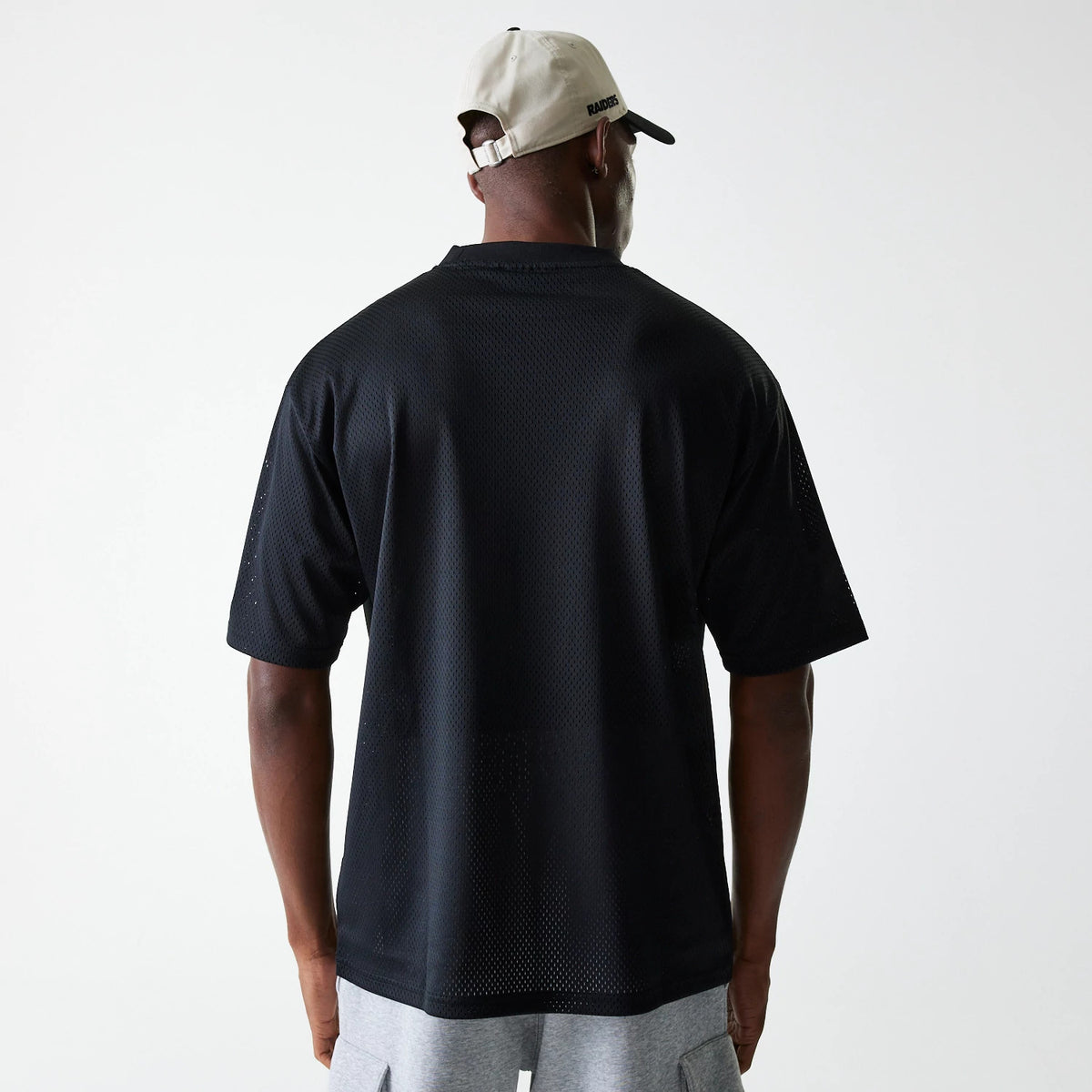 New Era Las Vegas Raiders NFL Essentials Black Oversized Mesh T-Shirt, rear view showing the micro mesh texture in black with short sleeves and a loose fit, worn by a person also wearing a beige cap with a raised logo on the back.
