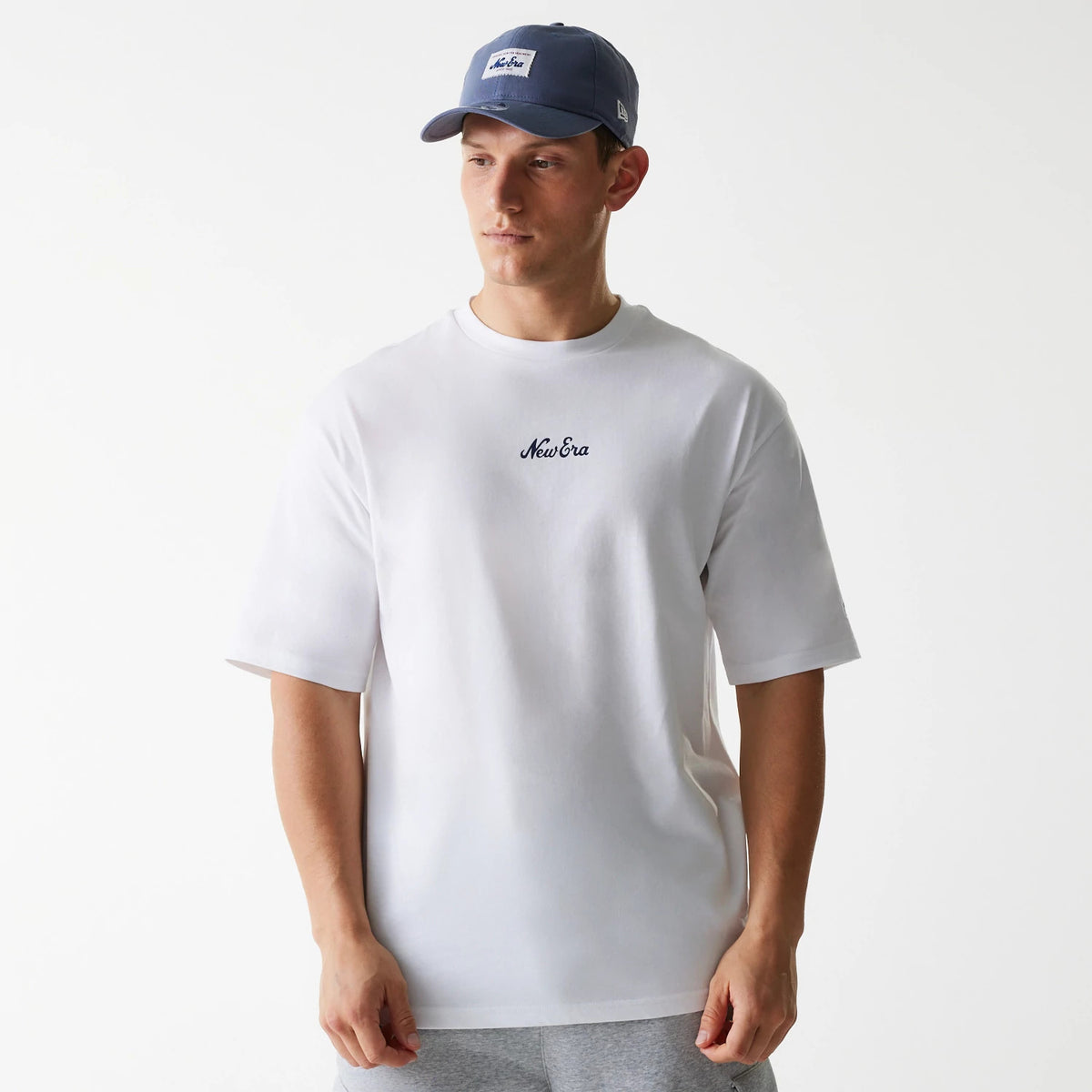 New Era Trucker Patch White Oversized T-Shirt, on-model view featuring a relaxed fit in white with a raised New Era logo on the chest, a blue cap with a patch on the front worn by the model, and grey trousers completing the look.