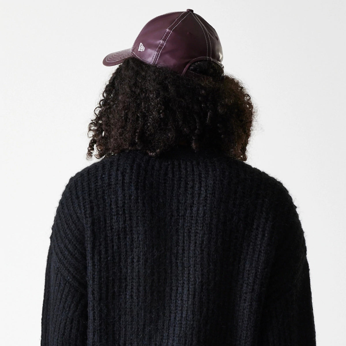 New Era New York Yankees Womens MLB Knit Black Cardigan, rear view on a model with curly hair wearing a black cardigan and a burgundy cap, showcasing the textured knit design and the New Era flag logo on the cap's left side.