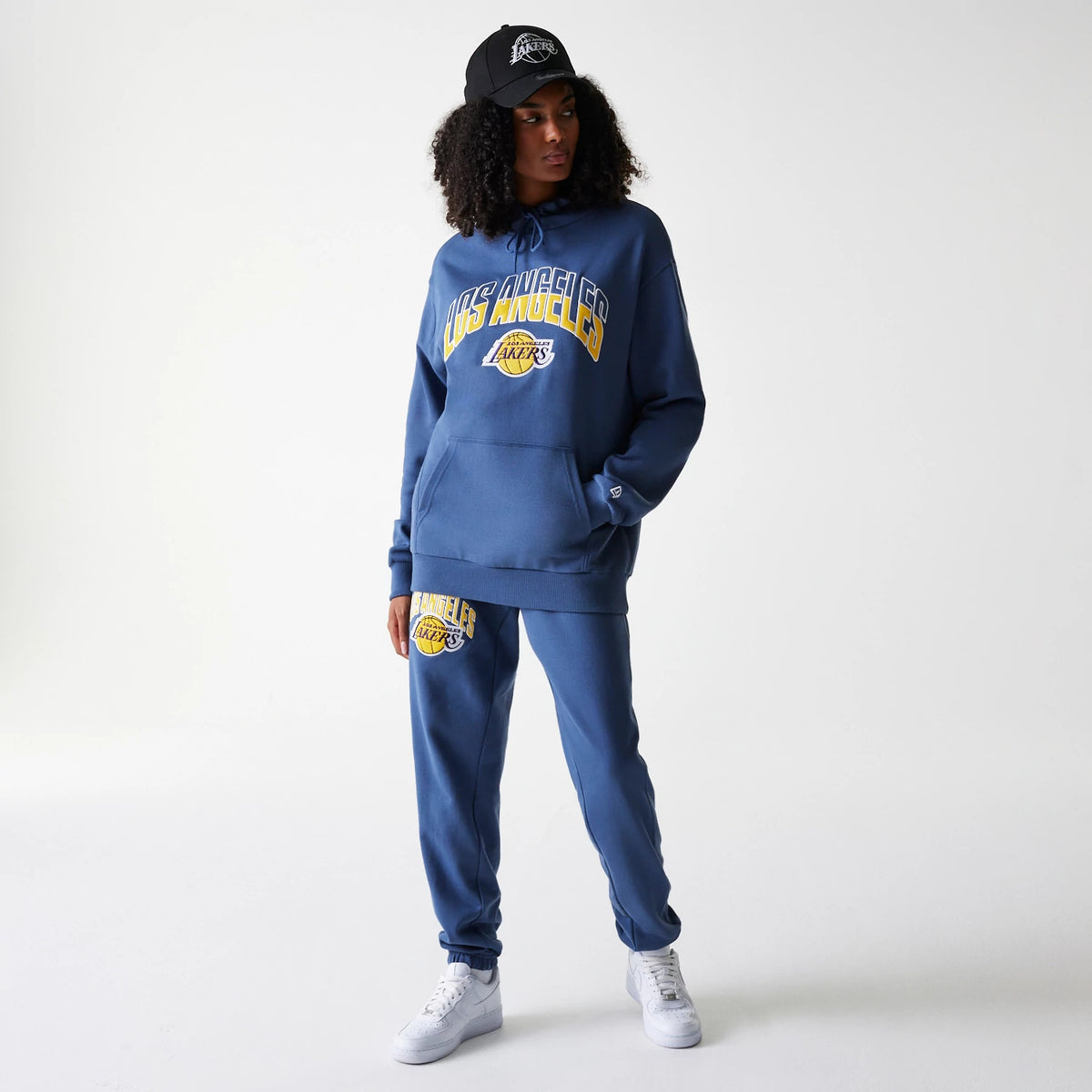 New Era LA Lakers NBA Arch Graphic Dark Blue Joggers, on a model with matching dark blue hoodie and cap, featuring a large Lakers logo on the left leg above the cuff and pockets on each side.