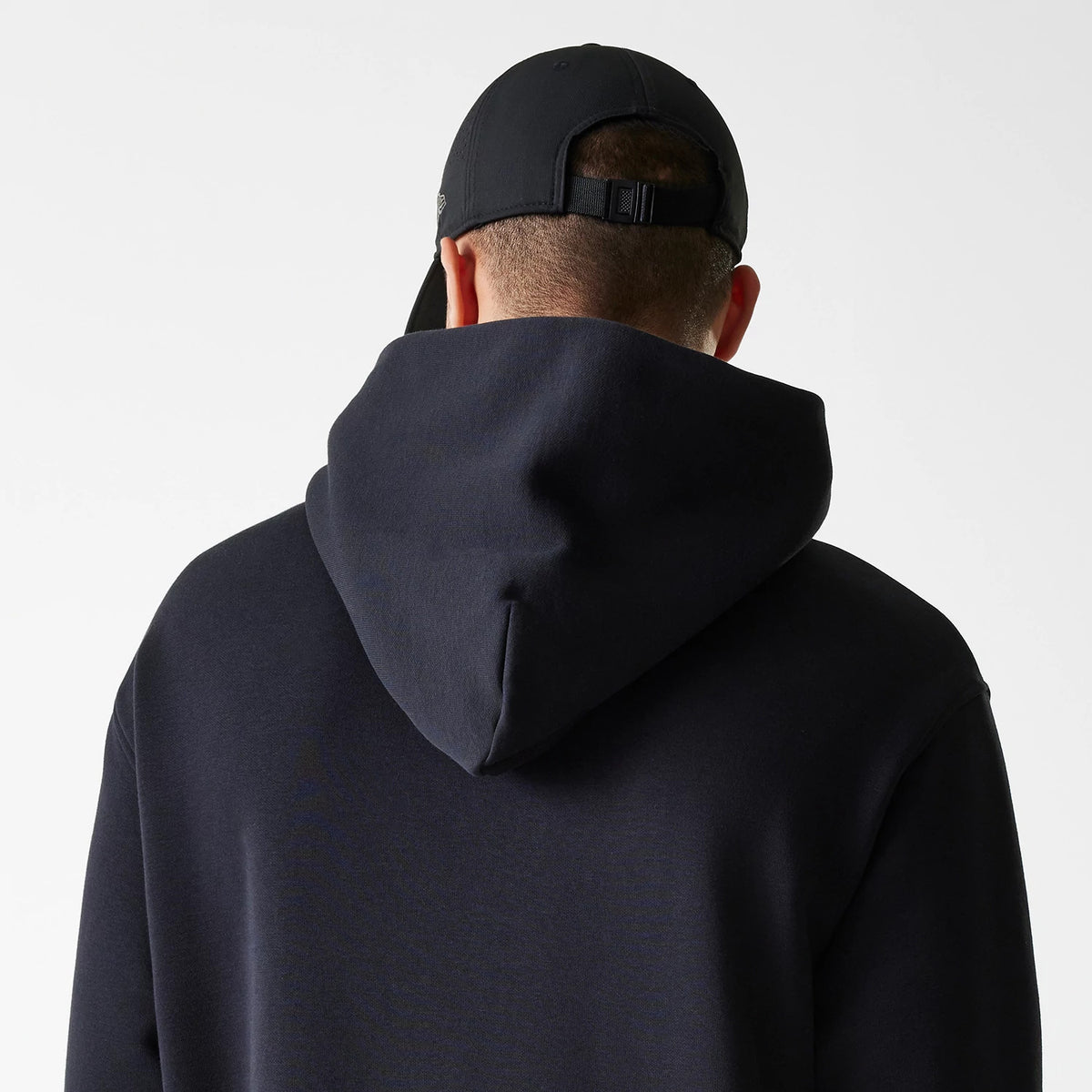 New Era Tech Black on Black Oversized Pullover Hoodie, rear view showing the oversized hood draping over shoulders, with a black debossed New Era logo on a black cap worn by the model.