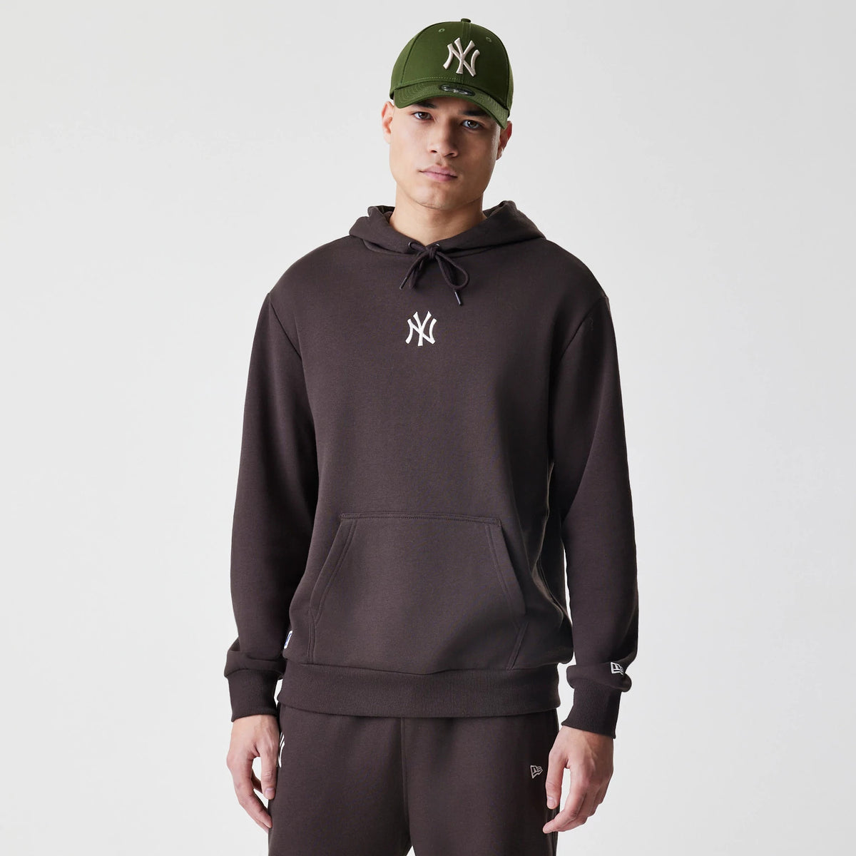 New Era New York Yankees MLB Midi League Essential Dark Brown Oversized Pullover Hoodie, front view on a model with matching dark trousers and a green cap, showing an embroidered white Yankees logo at the centre of the chest.
