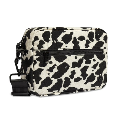 New Era Cow Jacquard Light Beige Waist Bag, side view showing the black zip pocket and clip buckle on the cow print pattern, with an embroidered New Era logo near the base on the front panel.