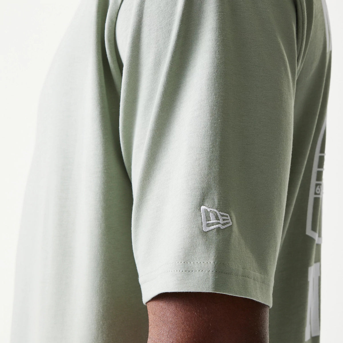 New Era Graphic Pastel Green Oversized T-Shirt, side view showing the light green sleeve with an embroidered white New Era logo near the cuff and part of a graphic design along the side.