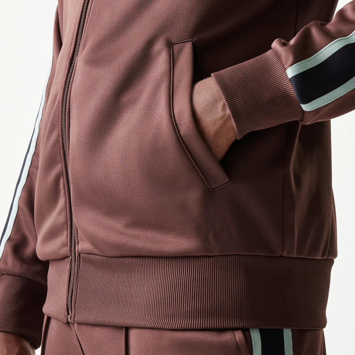 New Era Script Brown Track Jacket, front view with a focus on the zip closure and side pocket, showing ribbed cuffs and hem along with a model's hand resting in the pocket and striped detail on the sleeve.