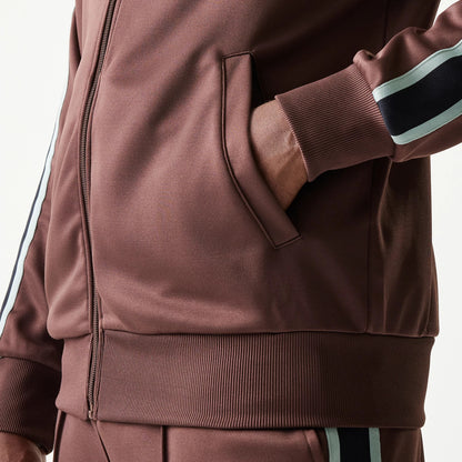 New Era Script Brown Track Jacket, front view with a focus on the zip closure and side pocket, showing ribbed cuffs and hem along with a model's hand resting in the pocket and striped detail on the sleeve.
